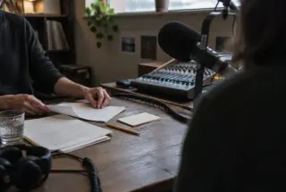 Hands organizing interview notes beside a microphone and audio console in a softly lit independent radio studio.