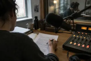 Radio producer revising a concise news script while a colleague listens behind the studio glass.